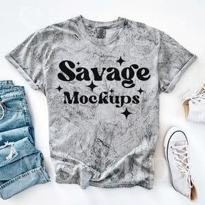 Peut inclure: Un t-shirt gris avec un aspect vieilli, sur lequel est inscrit "Savage Mockups" en noir avec des étoiles.