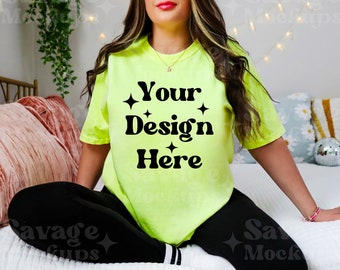 Op de afbeelding: Een vrouw draagt een fel neon groen T-shirt met de tekst "Your Design Here" in zwart. Het shirt is losjes gesneden en heeft een ronde hals. De vrouw zit op een bed met een roze kussen en een wit dekbed.