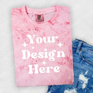 May include: Pink tie-dye t-shirt with the text "Your Design Here" in white. The shirt is folded and laid on a white surface with a pair of blue jeans.