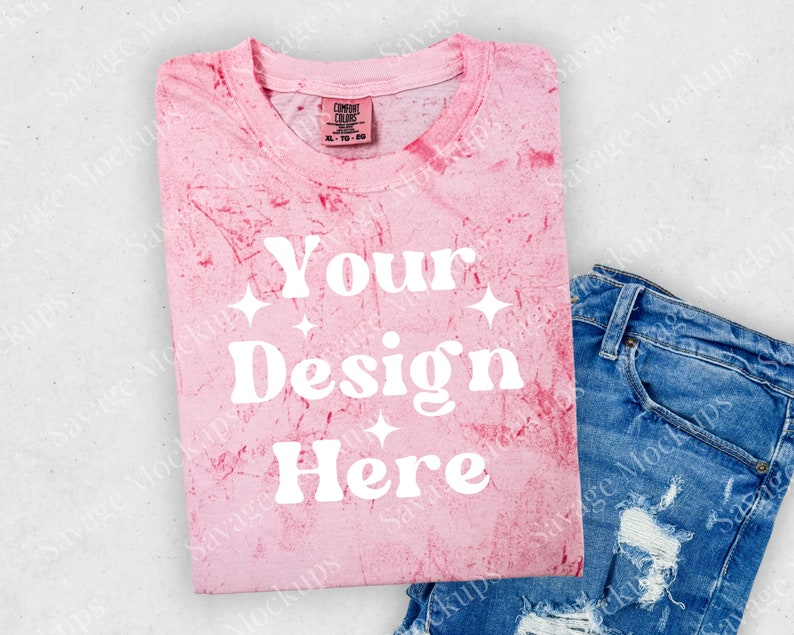 May include: Pink tie-dye t-shirt with the text "Your Design Here" in white. The shirt is folded and laid on a white surface with a pair of blue jeans.