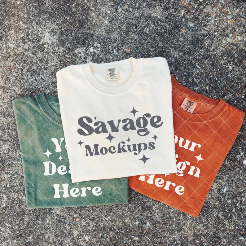 Pode incluir: Tr&ecirc;s camisetas dobradas em creme, verde e laranja. A camiseta creme apresenta o texto "Savage Mockups" com detalhes de estrelas. A camiseta verde diz "Your Design Here", e a camiseta laranja diz "Your Design Here".