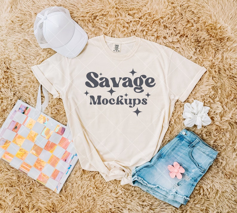 May include: A beige t-shirt with the text "Savage Mockups" in a retro font. The shirt is laid out on a fluffy beige rug with a white hat, a white mesh bag, a pair of denim shorts, and a white scrunchie.