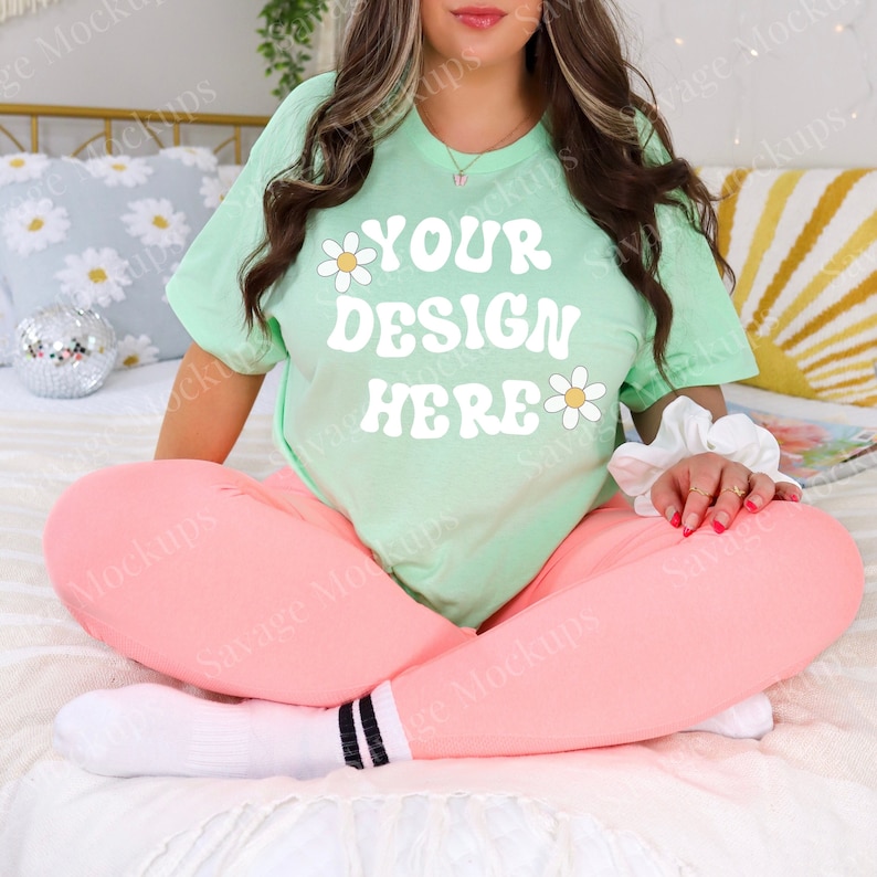 Puede incluir: Una camiseta verde claro de manga corta con un gr&aacute;fico blanco que dice "YOUR DESIGN HERE" con flores de margarita a cada lado del texto. La modelo lleva la camiseta con leggings rosas y calcetines blancos con rayas negras.