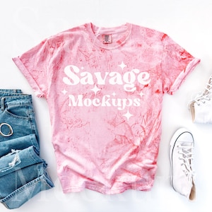 May include: A pink tie-dye t-shirt with the text "Savage Mockups" in white. The shirt is laid flat on a white surface with a pair of white high-top sneakers and a pair of blue jeans.