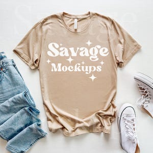 May include: A tan t-shirt with the text "Savage Mockups" in a retro font, accompanied by light blue jeans and white high-top sneakers. The items are arranged on a white surface.