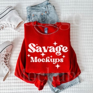 May include: Red sweatshirt with the text "Savage Mockups" in white. The sweatshirt is laid out on a white surface with a pair of white sneakers and blue jeans.