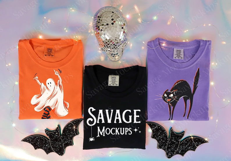 May include: Three Halloween themed t-shirts with different designs. The first shirt is orange with a white ghost dancing. The second shirt is black with the text "SAVAGE MOCKUPS" in white. The third shirt is purple with a black cat. All three shirts are folded and laid out on a light blue background with a disco ball and string lights.