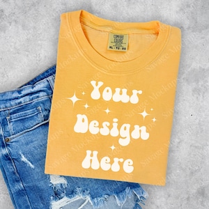May include: A yellow t-shirt with a white retro-style graphic that says "Your Design Here" and a few white stars. The t-shirt is folded and placed on top of a pair of blue denim jeans.