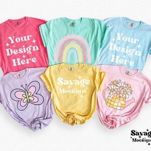May include: Five different colored t-shirts with various designs. The shirts are laid out on a white surface. The designs include a rainbow, a butterfly, a watering can with flowers, and the text "Your Design Here" and "Savage Mockups".
