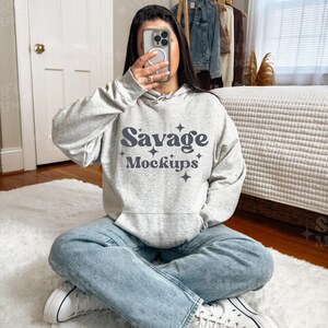 May include: A woman wearing a gray hoodie with the text "Savage Mockups" in a retro font. She is sitting on a white fluffy rug with her legs crossed and wearing white sneakers.