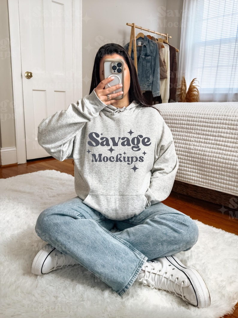 May include: A woman wearing a gray hoodie with the text "Savage Mockups" in a retro font. She is sitting on a white fluffy rug with her legs crossed and wearing white sneakers.