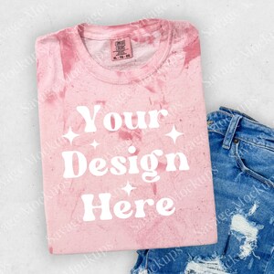 May include: Pink tie-dye t-shirt with the text "Your Design Here" in white. The shirt is folded and laid on a white surface with a pair of blue jeans.
