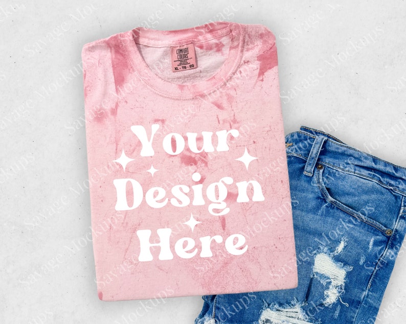 May include: Pink tie-dye t-shirt with the text "Your Design Here" in white. The shirt is folded and laid on a white surface with a pair of blue jeans.