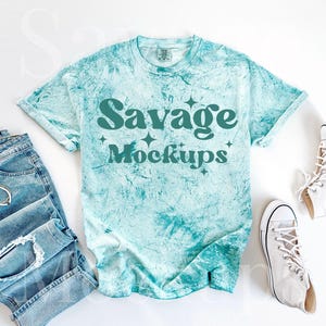 May include: A turquoise blue tie-dye t-shirt with the text "Savage Mockups" in teal green. The t-shirt is laid flat on a white surface with a pair of white high-top sneakers and a pair of blue jeans.
