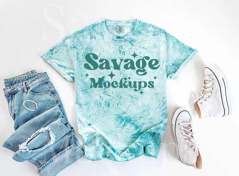 May include: A turquoise blue tie-dye t-shirt with the text "Savage Mockups" in teal green. The t-shirt is laid flat on a white surface with a pair of white high-top sneakers and a pair of blue jeans.