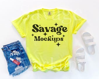 Op de afbeelding: Een felgeel T-shirt met de tekst "Savage Mockups" in zwarte letters. Het shirt ligt plat op een wit oppervlak.