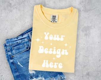 May include: A pale yellow t-shirt with a white graphic that says "Your Design Here" in a retro font. The shirt is folded and laid on top of a pair of blue denim shorts.