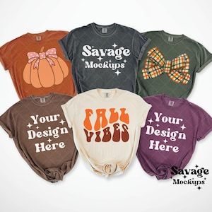 Puede incluir: Se muestran seis camisetas en varios colores, incluyendo marrón, naranja y morado. Cada camiseta presenta un diseño único, como una calabaza, un lazo o los textos "Fall Vibes" y "Your Design Here". Las camisetas se exhiben sobre un fondo blanco.