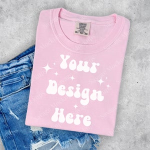 May include: A pink t-shirt with the text "Your Design Here" in a retro font. The shirt is folded and placed on top of a pair of blue jeans.
