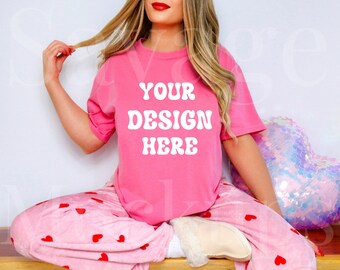 May include: A pink t-shirt with the text "YOUR DESIGN HERE" in white. The shirt is being worn by a person sitting on a wooden surface with a pink heart-shaped sequin pillow.