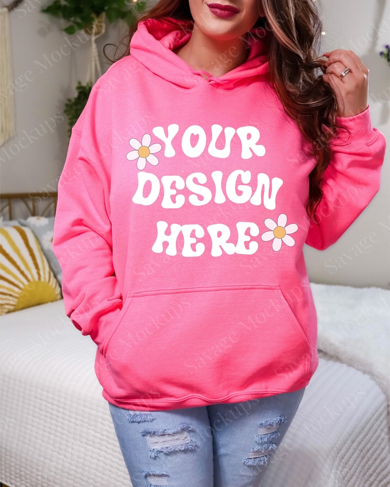 May include: A bright pink hoodie with the text "YOUR DESIGN HERE" in white, accompanied by daisy graphics. The hoodie has a kangaroo pocket and a drawstring hood. The model is wearing light blue jeans.