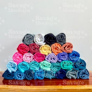 May include: A stack of rolled up t-shirts in various colors, including pink, blue, green, yellow, and orange. The shirts are arranged in a pyramid shape on a wooden surface.