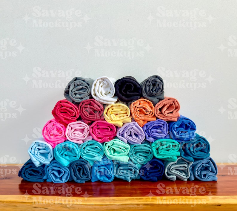 May include: A stack of rolled up t-shirts in various colors, including pink, blue, green, yellow, and orange. The shirts are arranged in a pyramid shape on a wooden surface.