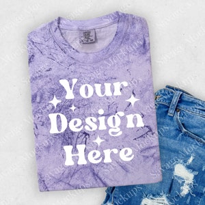 May include: A purple tie-dye t-shirt with the text "Your Design Here" in white. The shirt is folded and placed on a white surface with a pair of blue jeans.