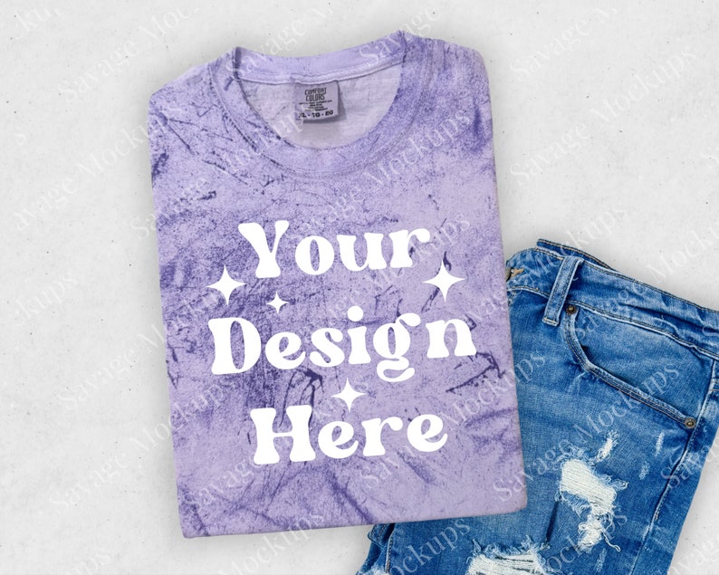 May include: A purple tie-dye t-shirt with the text "Your Design Here" in white. The shirt is folded and placed on a white surface with a pair of blue jeans.