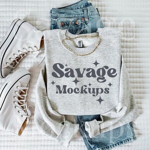 May include: A gray sweatshirt with the text "Savage Mockups" in a retro font. The sweatshirt is laid out on a white surface with a pair of white sneakers and blue jeans.