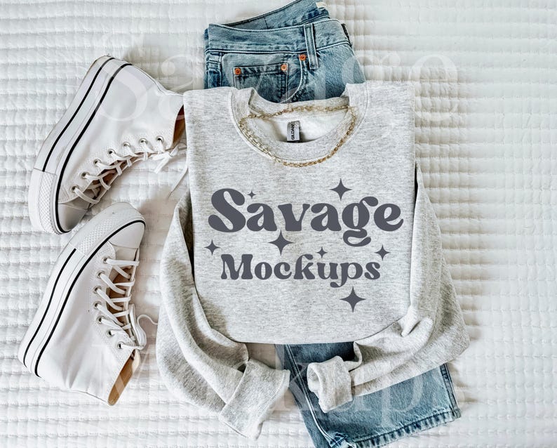 May include: A gray sweatshirt with the text "Savage Mockups" in a retro font. The sweatshirt is laid out on a white surface with a pair of white sneakers and blue jeans.