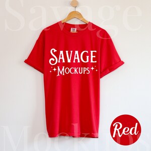 May include: Red t-shirt with the text "SAVAGE MOCKUPS" in white on the front. The shirt is hanging on a wooden hanger against a white wall.