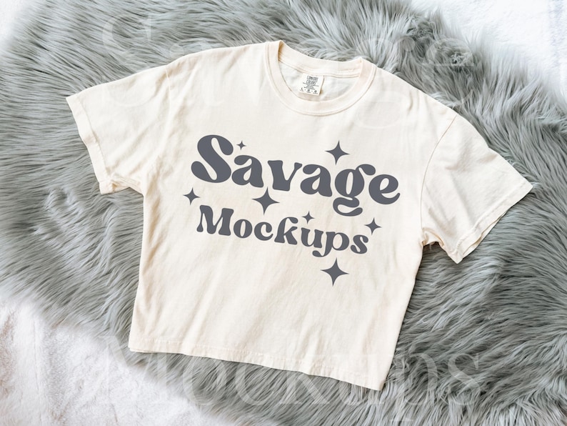 May include: A cream colored t-shirt with the text "Savage Mockups" in a dark gray font with stars around the text.