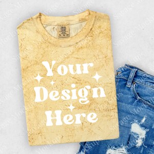 May include: A folded yellow t-shirt with a distressed look. The shirt has the text "Your Design Here" in white. The shirt is laying on a light gray surface with a pair of blue jeans with distressed details.