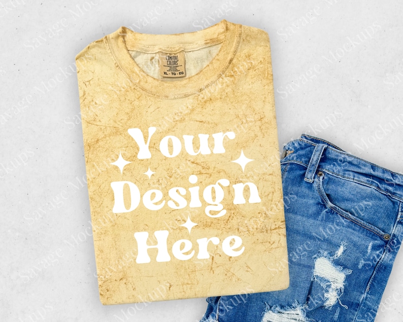 May include: A folded yellow t-shirt with a distressed look. The shirt has the text "Your Design Here" in white. The shirt is laying on a light gray surface with a pair of blue jeans with distressed details.