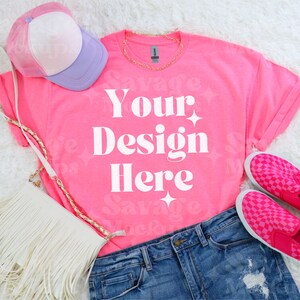 Puede incluir: Una camiseta rosa con el texto "Your Design Here" en blanco. La camiseta está colocada sobre una superficie blanca y peluda con un bolso blanco, una gorra de béisbol rosa y morada y zapatos de tela rosa a cuadros.