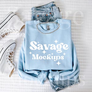 May include: A light blue sweatshirt with the text "Savage Mockups" in white. The sweatshirt is laid out on a white surface with a pair of white high-top trainers and blue jeans.