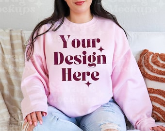 Könnte beinhalten: Ein rosa Sweatshirt mit dem Text "Your Design Here" in maroonfarbener Schrift. Das Sweatshirt wird von einer Person getragen, die auf einem Sofa sitzt.
