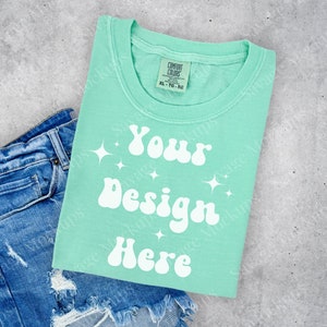 May include: A light green t-shirt with the text "Your Design Here" in white, with a retro font and sparkling stars. The t-shirt is folded and placed on top of a pair of blue denim shorts.