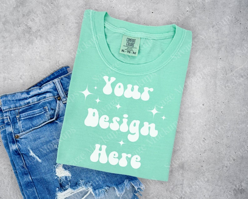 May include: A light green t-shirt with the text "Your Design Here" in white, with a retro font and sparkling stars. The t-shirt is folded and placed on top of a pair of blue denim shorts.