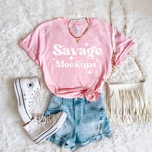 May include: A pink t-shirt with the text "Savage Mockups" in white. The shirt is laying on a white fuzzy surface with a pair of white high-top sneakers, a pair of light blue denim shorts, and a white fringed purse.