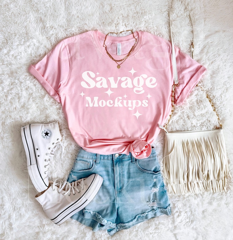 May include: A pink t-shirt with the text "Savage Mockups" in white. The shirt is laying on a white fuzzy surface with a pair of white high-top sneakers, a pair of light blue denim shorts, and a white fringed purse.
