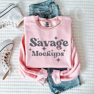May include: A pink sweatshirt with the text "Savage Mockups" in a retro font. The sweatshirt is laid out on a white surface with a pair of white sneakers and blue jeans.