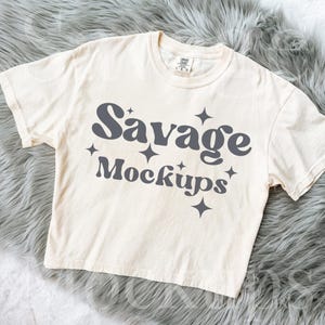 May include: Cream-colored t-shirt with the text "Savage Mockups" in dark gray, accented with star graphics. The shirt is laid on a fluffy, gray faux fur surface. A casual, unisex tee.