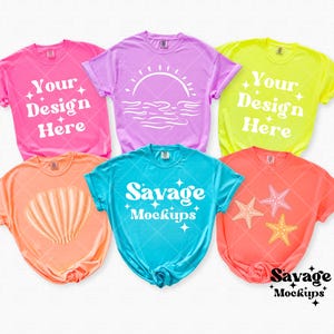 May include: Five different colored t-shirts with various designs. The shirts are laid out flat on a white surface. The designs include a seashell, a sunset, two starfish, and the text "Your Design Here" and "Savage Mockups".