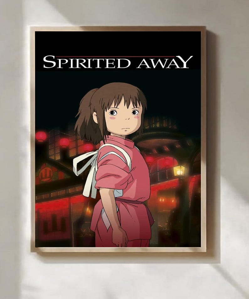 Op de afbeelding: Een ingelijste print met een personage uit de animatiefilm "Spirited Away". Het kunstwerk toont een jong meisje in een roze outfit met een witte band, tegen een achtergrond van een nachtelijk stadsbeeld met rode lantaarns. De titel "SPIRITED AWAY" staat bovenaan.