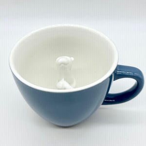 May include: A blue ceramic coffee mug with a white interior and a white sculpted bear inside. The mug has a curved shape and a handle on the side. The background is plain white.