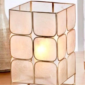 May include: A square decorative lamp with a gold metal frame and off-white, square-shaped panels. The lamp is lit from within, creating a warm glow. The lamp is sitting on a wooden surface.