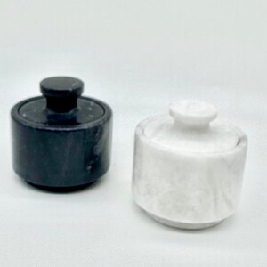 May include: Two small cylindrical containers with lids, one crafted from solid black stone and the other from solid white stone. Each container features a smooth, polished finish and a small, circular knob handle centered on the top of the lid.