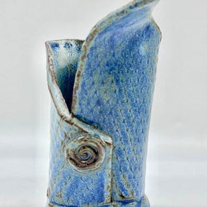 May include: A cylindrical ceramic vase with a textured blue glaze featuring a cross-hatched pattern. The vase has an asymmetrical, folded-over rim and a prominent spiral-shaped decorative element on the front, with brown edges along the seams.